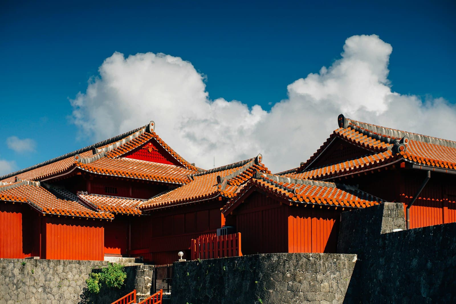 A traditional Okinawan/Ryukyuan temple or fortified compound with characteristic orange-tiled curved roofs, red wooden walls and doors, black trim, and gray stone perimeter walls under a clear blue sky. - Photo by WS Chae on Unsplash