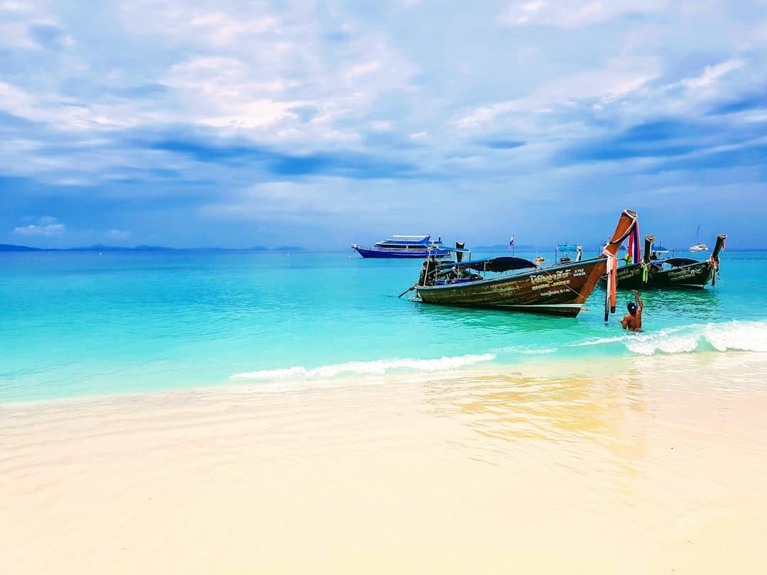 A pristine tropical beach with crystal-clear turquoise waters, white sand, and traditional Thai longtail boats anchored in shallow waters, with a larger tour boat visible in the background. - Photo by The DK Photography on Unsplash