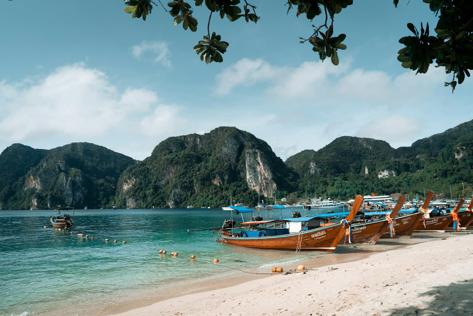 A scenic beach cove with orange and blue wooden longtail boats moored in turquoise water, dramatic limestone cliffs in the background, and white sand beach with tropical foliage overhead. - Photo by Vaskar Sam on Unsplash