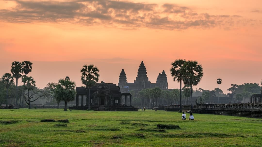 Angkor Wat temple complex photographed at sunrise from the western approach, with the iconic central towers silhouetted against an orange sky, palm trees framing the view, and visitors on the main courtyard. - Photo by Roth Chanvirak on Unsplash