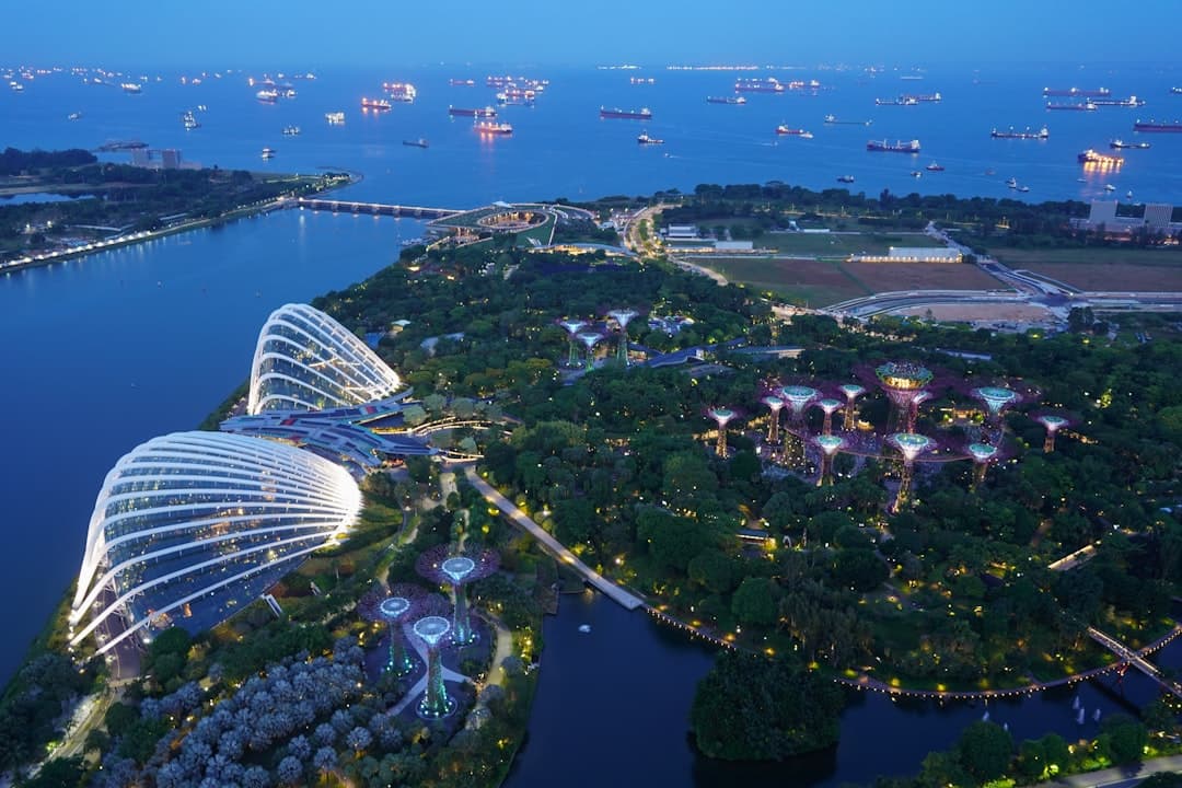 Aerial twilight photograph of Marina Bay in Singapore showing the illuminated Marina Bay Sands hotel, Gardens by the Bay with its lit Supertrees, and numerous cargo ships anchored in the harbor. - Photo by Zion C on Unsplash