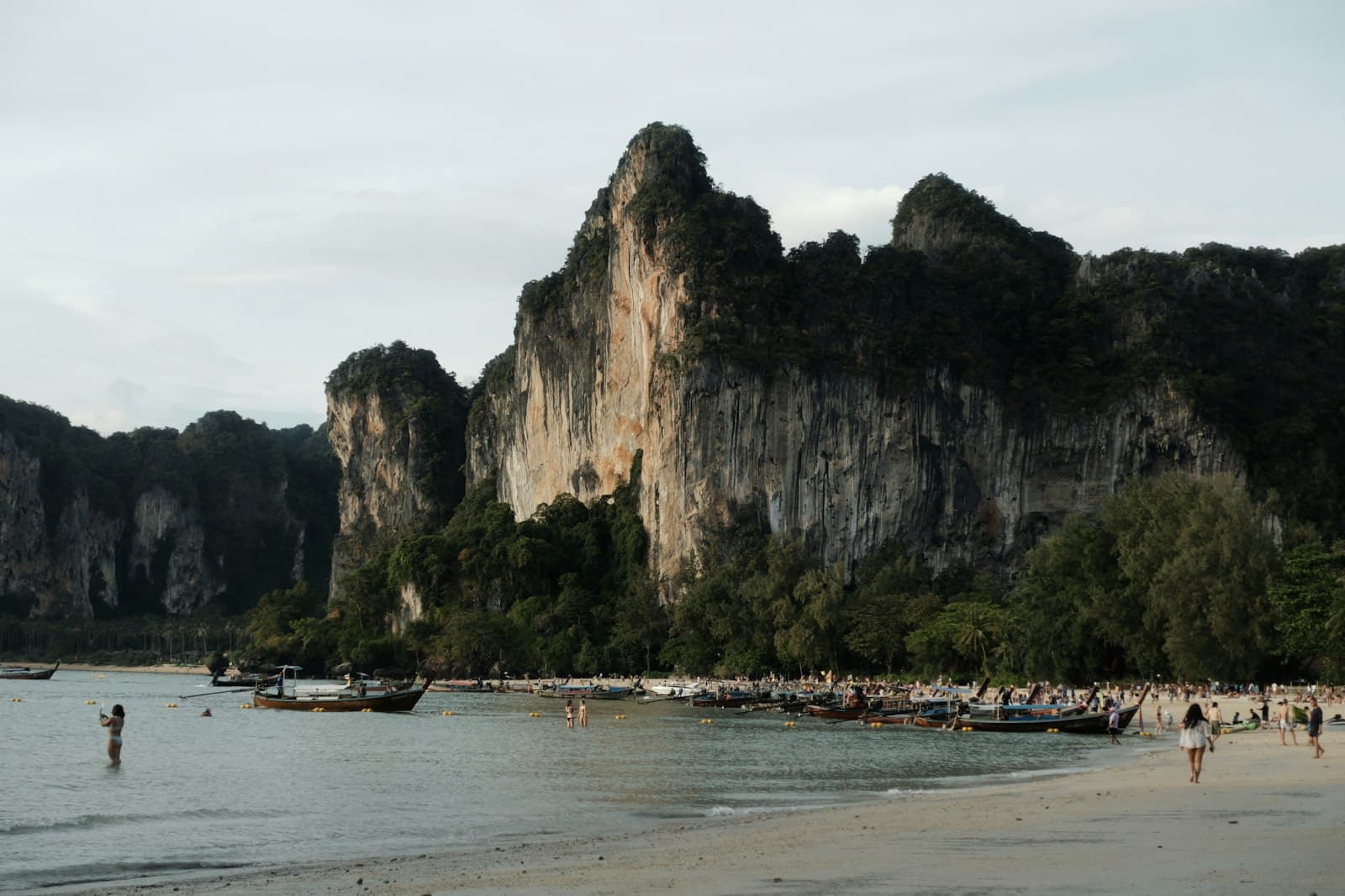 A busy tropical beach backed by dramatic limestone karst cliffs, with tourists swimming and sunbathing, long-tail boats moored in shallow turquoise water, and dense jungle vegetation covering the cliff faces. - Photo by SERGEI BEZZUBOV on Unsplash