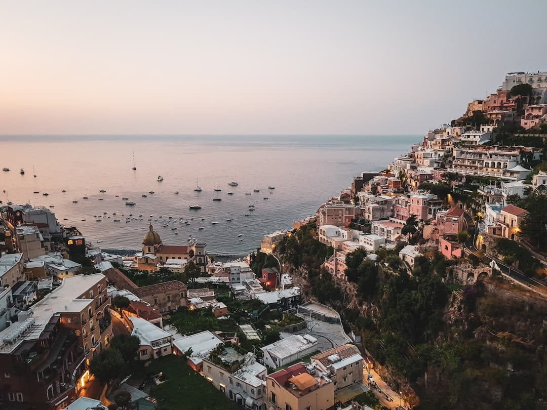 Golden hour photograph of the iconic clifftop village of Positano cascading down to the Tyrrhenian Sea, with colorful pastel buildings stacked vertically on the hillside, a church dome, and numerous small boats anchored in the calm bay below. - Photo by Dimitry B on Unsplash