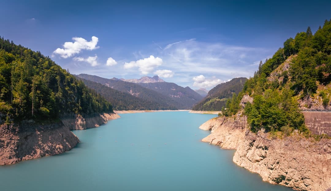 A stunning turquoise Alpine reservoir surrounded by steep forested mountainsides, limestone cliffs, and a visible dam structure, photographed on a clear day with cumulus clouds. - Photo by Christian Lendl on Unsplash