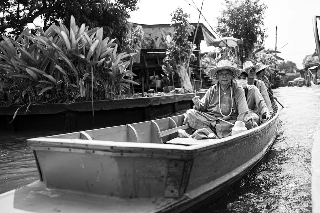 Black and white photograph of boatmen in traditional conical hats navigating a wooden boat past riverside dwellings and tropical vegetation along what appears to be the Mekong River or a major tributary. - Photo by Leo_Visions on Unsplash