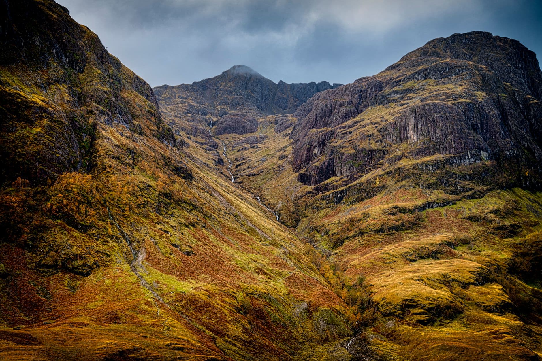 A dramatic Highland valley with steep moss-covered slopes, columnar basalt cliffs, and a central burn, viewed looking up toward a distant peak under partly cloudy skies. - Photo by Robert Hacker on Pexels