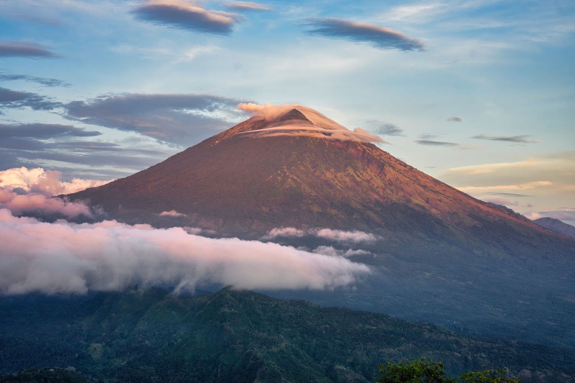 A conical stratovolcano at sunset with its peak illuminated in golden light, partially shrouded in clouds at its base, surrounded by tropical forest. - Photo by Tom Fisk on Pexels