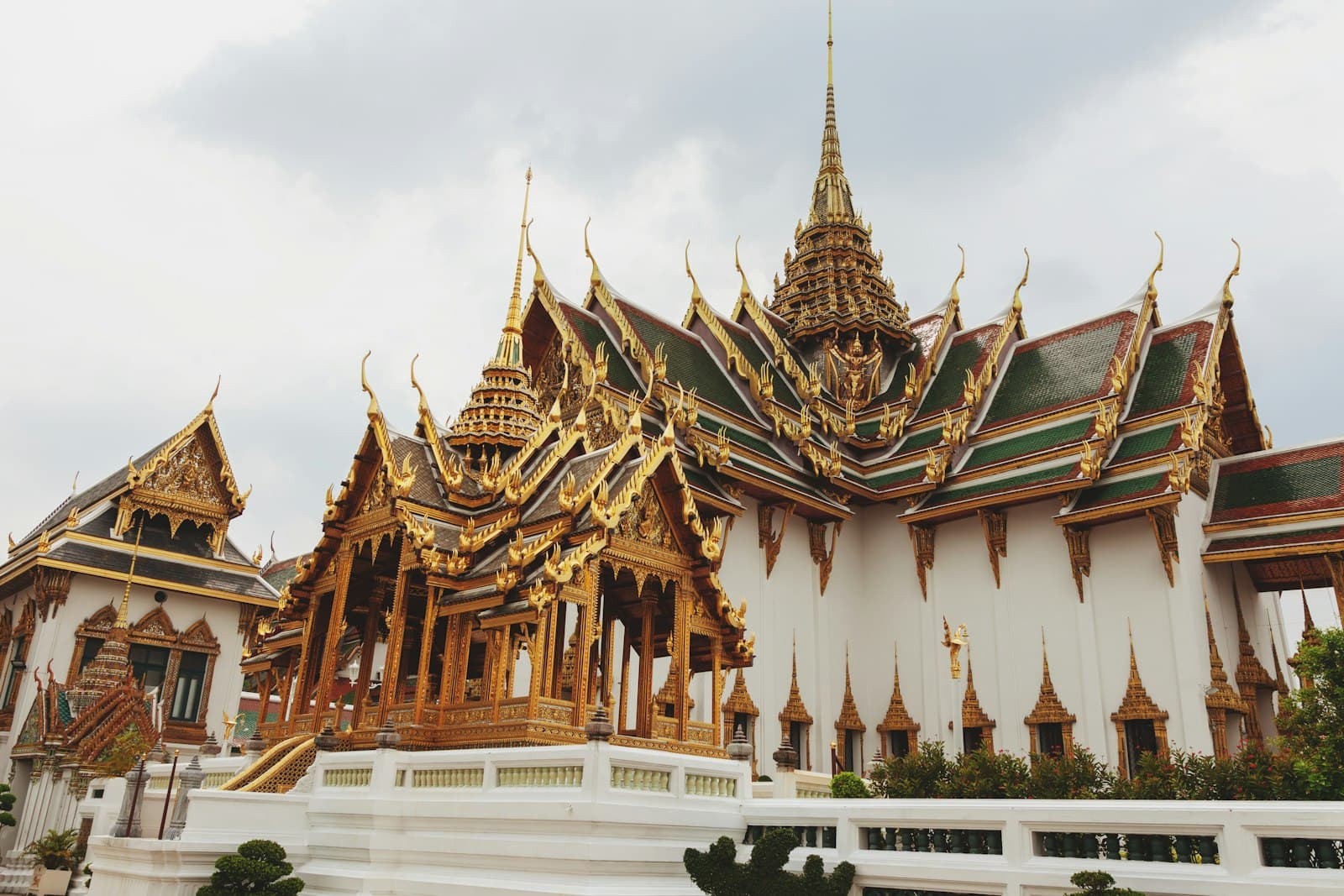 A grand Thai Buddhist temple with multiple golden-spired buildings, ornate wooden decorative elements, white marble railings, and characteristic Southeast Asian religious architecture under an overcast sky. - Photo by Rowan Heuvel on Unsplash