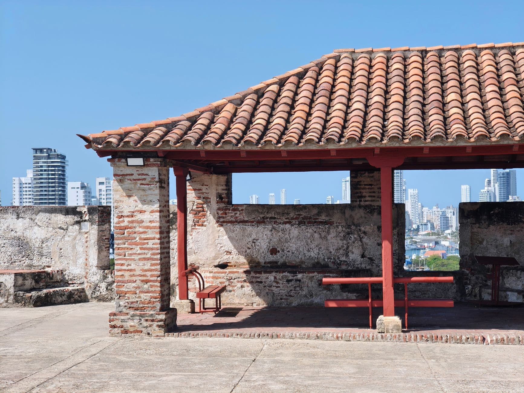A restored colonial-era structure with terracotta roof tiles and red-painted wooden supports overlooks modern Cartagena's skyline from what appears to be the city's historic fortified walls. - Photo by Woody Willis on Pexels