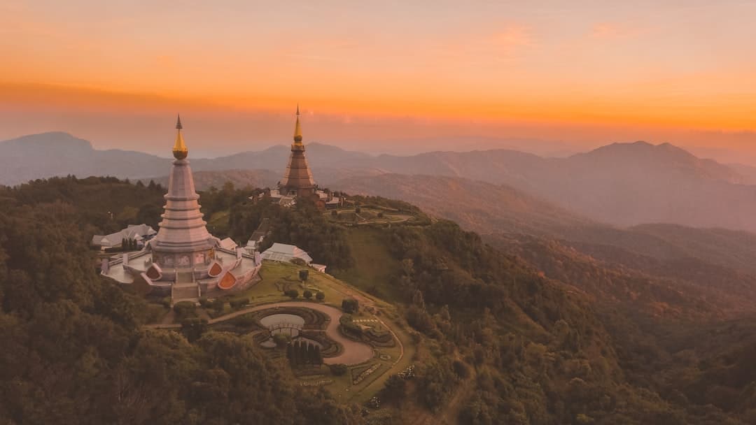 Aerial view of two illuminated Buddhist pagodas on forested mountain peaks at sunset or sunrise, with a sprawling mountain landscape and valley visible in the background. - Photo by Bharath Mohan on Unsplash