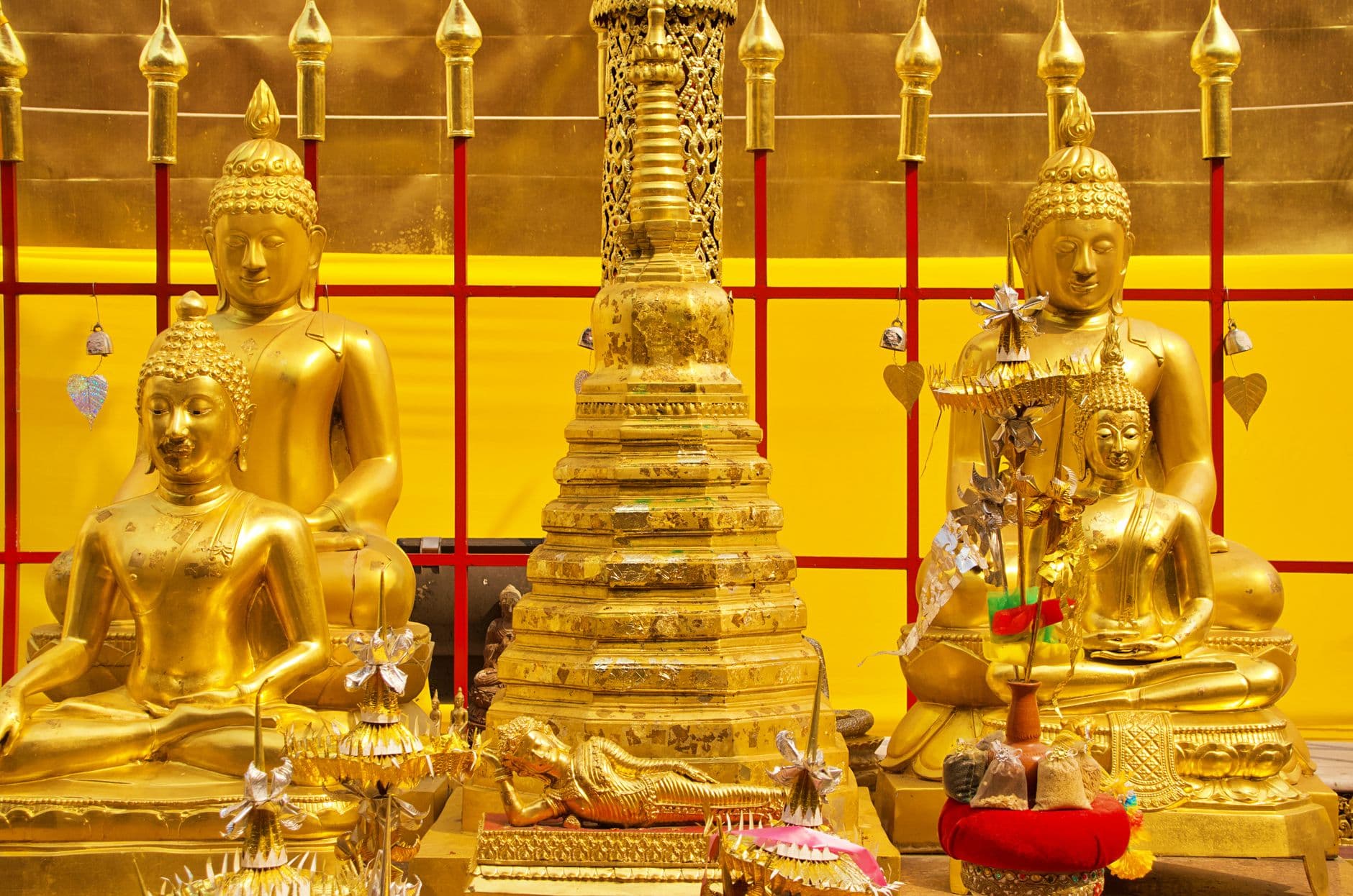 A richly decorated Buddhist shrine or temple interior displaying multiple golden Buddha statues in meditation posture, a central tiered stupa, ornamental spires, and fresh floral and ribbon offerings against a bright yellow wall with red cross-hatched framing. - Photo by Khan ishaan on Pexels