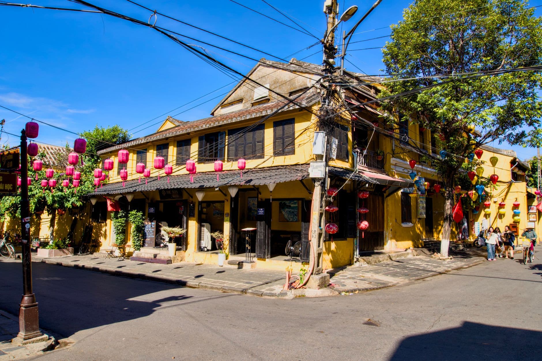 A street corner in Hoi An's Ancient Town featuring yellow French colonial buildings decorated with traditional pink silk lanterns, dark wooden shutters, and vine-covered facades. - Photo by Tuan Vy on Pexels