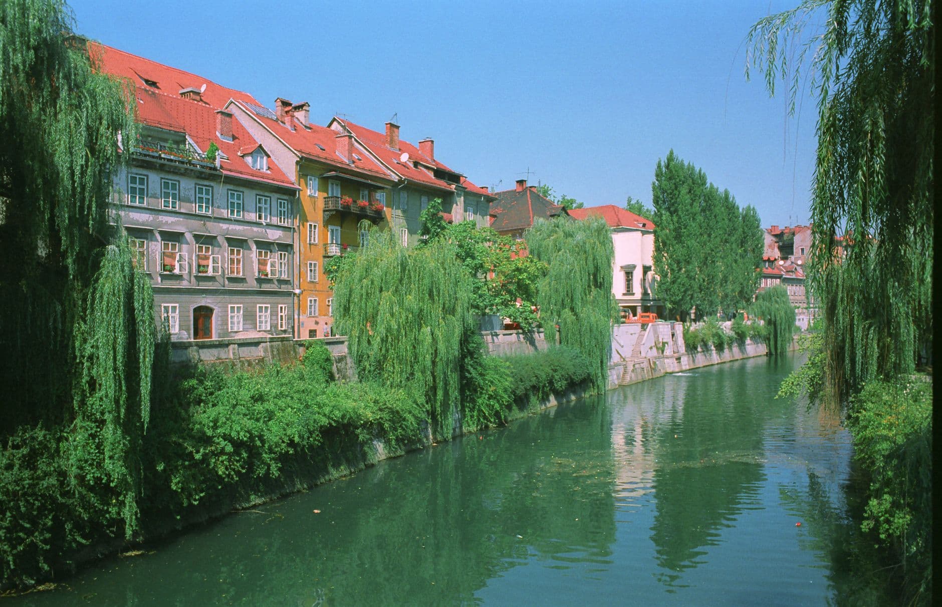 A scenic view down the Ljubljanica River through Ljubljana's Old Town, lined with colorful period buildings with red tile roofs and framed by weeping willow trees. - Photo by Carlo Primo on Pexels