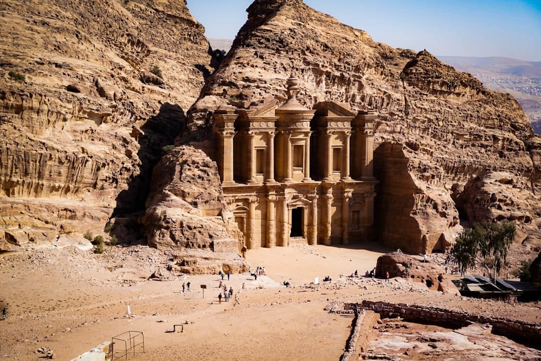 This photograph shows the Al-Khazneh (Treasury) at Petra, Jordan—an elaborate Nabatean carved facade hewn from rose-colored sandstone cliffs, with visitors visible at its base for scale. - Photo by Jameson Berrios on Unsplash