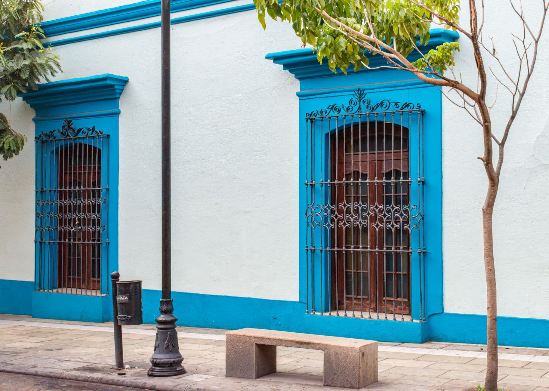 A colonial-era building facade with white-washed walls and bright turquoise trim, featuring two barred windows with decorative wrought-iron grillwork, street furniture, and tropical vegetation. - Photo by Jonathan Tesmaye (Kuns) on Pexels