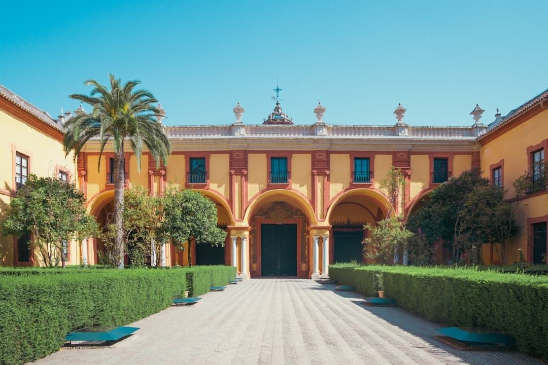 A grand courtyard of a colonial-era palace or mansion in Seville, featuring yellow ochre walls with red trim, Romanesque arches, manicured hedges, and a palm tree beneath clear blue sky. - Photo by Lianhao Qu on Unsplash