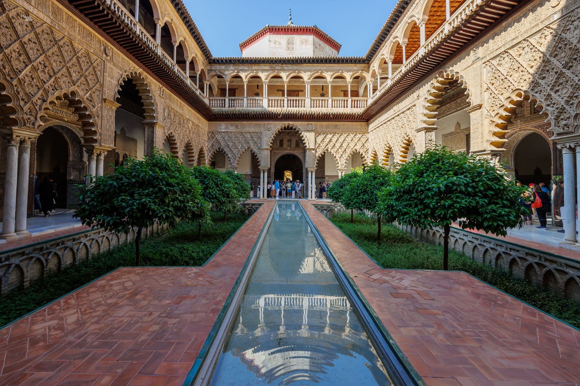 The Patio de las Doncellas (Court of the Maidens) in Seville's Real Alcázar, showing the characteristic Mudéjar architecture with decorative horseshoe arches, elaborate stone carvings, and a central water channel reflecting the surrounding arcades. - Photo by Igor Passchier on Pexels