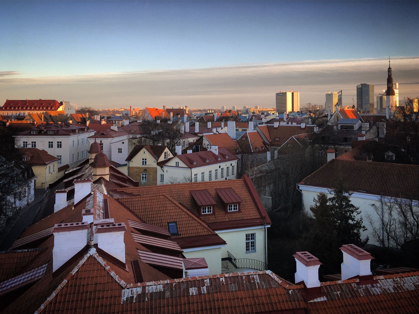 A panoramic view of Tallinn's Old Town rooftops at sunset, showing traditional red-tiled and white-walled medieval buildings with the city's modern skyline and church spires visible in the background. - Photo by Sergei Gussev on Pexels
