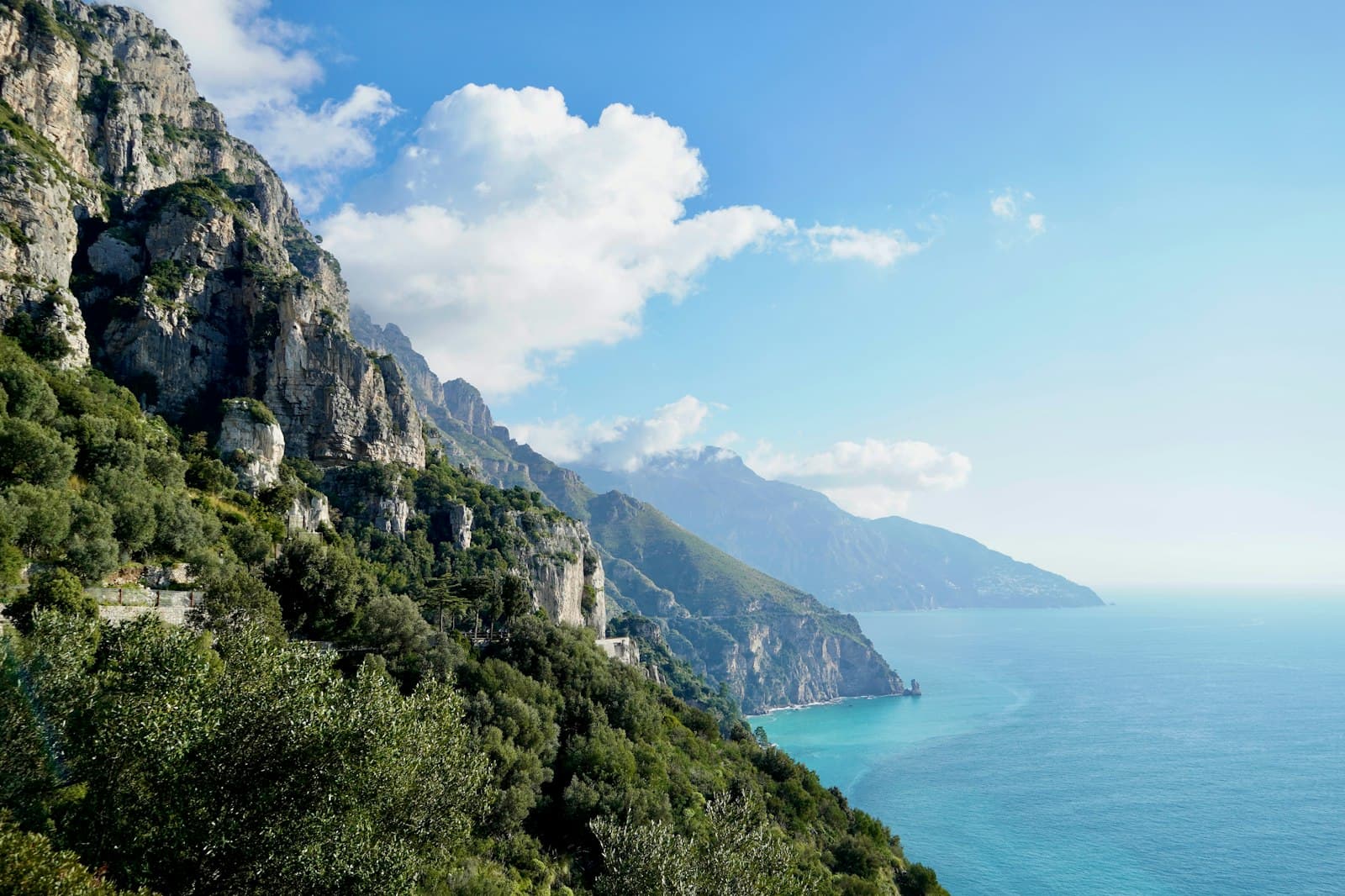 Steep limestone cliffs covered in Mediterranean vegetation rise above turquoise sea waters along a dramatic rocky coastline, viewed from a coastal road or path. - Photo by Meina Yin on Unsplash