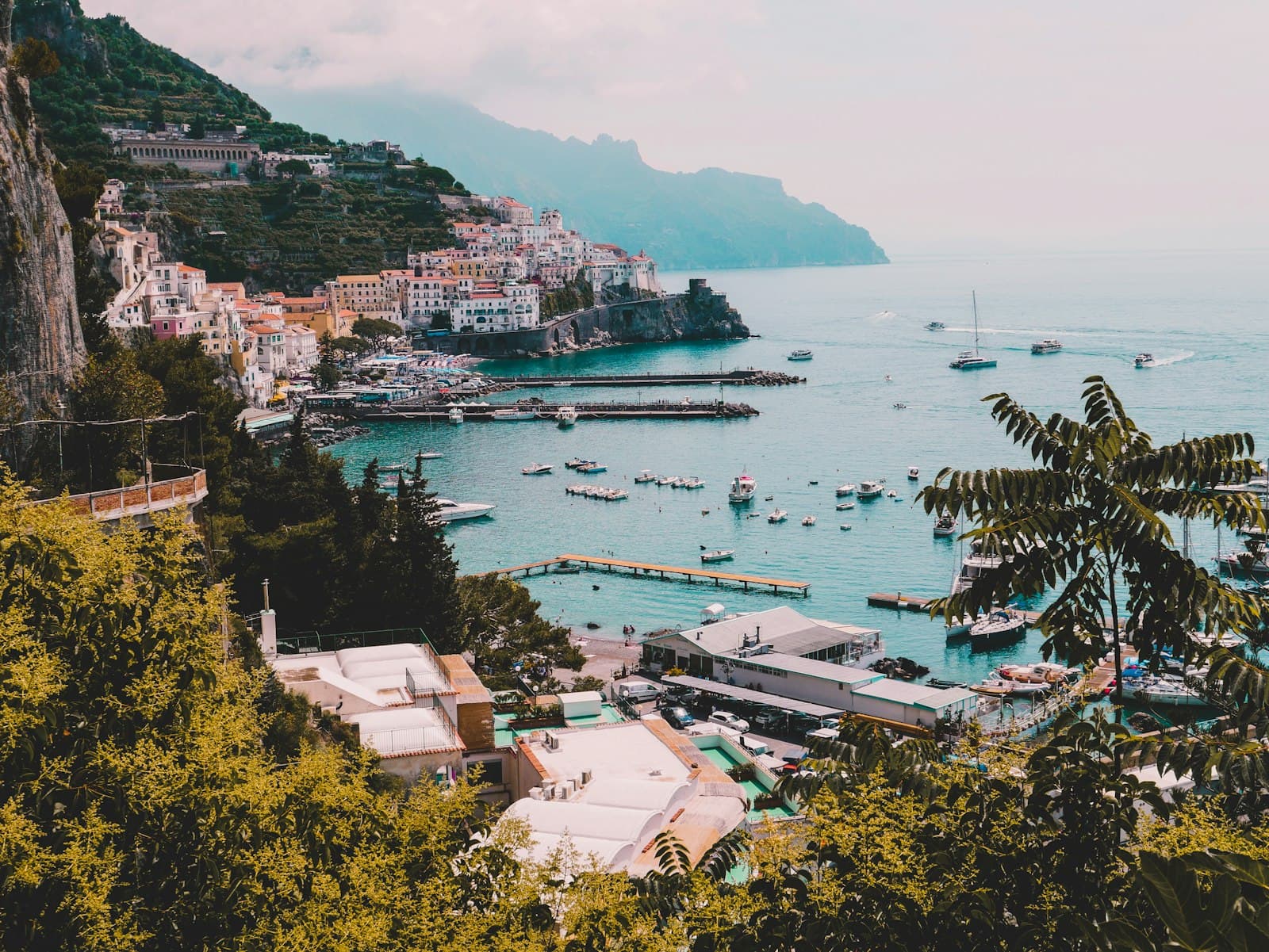 A scenic coastal town built vertically into a steep hillside overlooking a turquoise bay filled with moored boats, with pastel-colored buildings, lush vegetation, and dramatic cliff formations. - Photo by silvia trigo on Unsplash