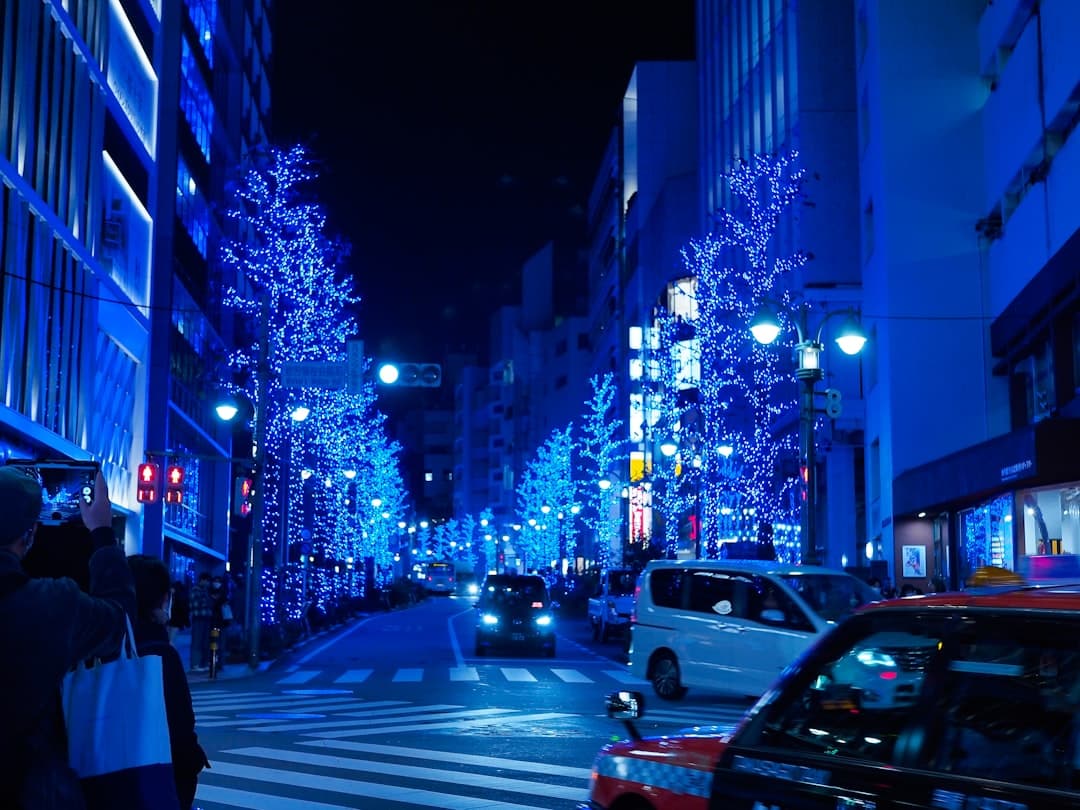 A nighttime urban street scene with modern buildings, illuminated blue decorative trees lining both sides of a pedestrian crossing, traffic, and characteristic Japanese city infrastructure. - Photo by Yuika Takamura on Unsplash