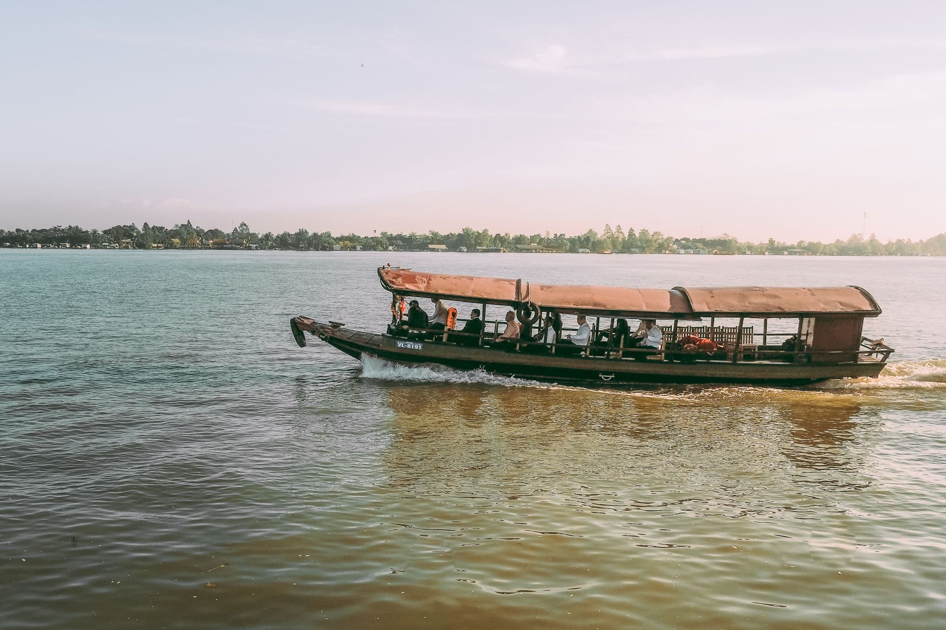 A wooden passenger ferry with a rust-colored canvas canopy carries tourists across a wide, shallow section of the Mekong River, with dense riverside vegetation visible in the distance. - Photo by Vũ Bụi on Pexels