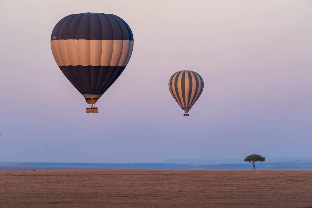 Two hot air balloons drift above the Maasai Mara savanna at dawn, with an iconic acacia tree and grazing wildlife visible on the plain below. - Photo by Boopathi Rajaa Nedunchezhiyan on Unsplash