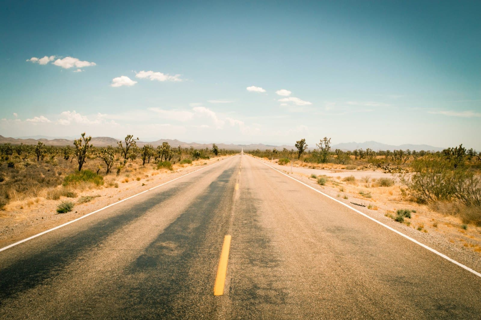 A straight desert highway with yellow and white road markings stretches toward distant mountains through a Mojave Desert landscape dotted with Joshua trees and sparse scrubland under clear blue sky. - Photo by Tyke Jones on Unsplash