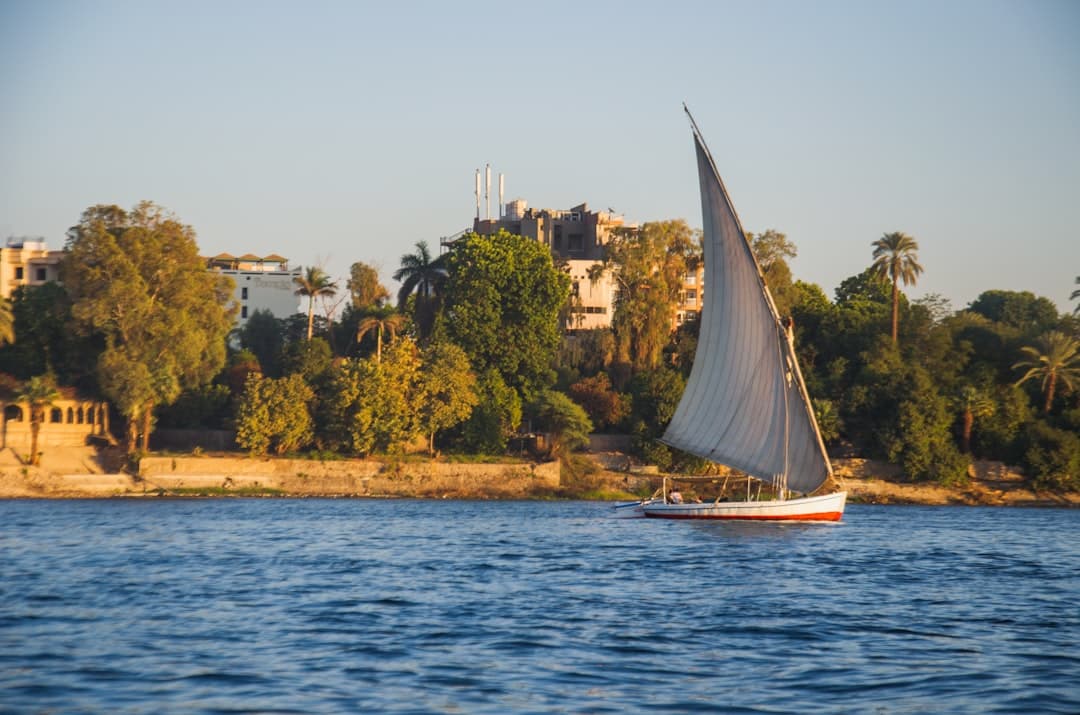 A traditional Egyptian felucca sailboat with blue-grey sail and red hull sails past colonial-era riverfront buildings on the Nile River, with palm trees and sandy banks visible in golden afternoon light. - Photo by Ale on Unsplash