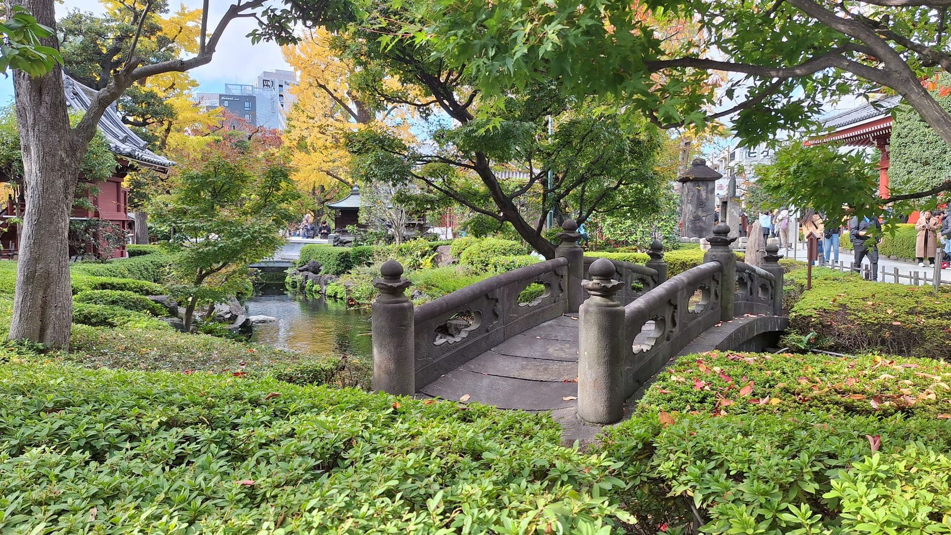 A serene Japanese temple or shrine garden featuring a traditional arched stone bridge over a small stream, surrounded by mature trees in autumn color transition, manicured hedges, and traditional architecture. - Photo by Suphot Punnachaiya on Pexels