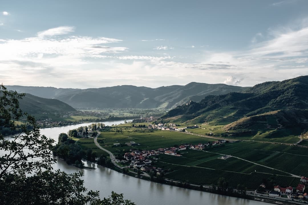 An elevated view of the Wachau Valley showing a dramatic meander in the Danube River surrounded by steep, forested hills, with vineyard terraces, small villages with red-roofed houses, and green pastures in the floodplain. - Photo by Thomas Peham on Unsplash