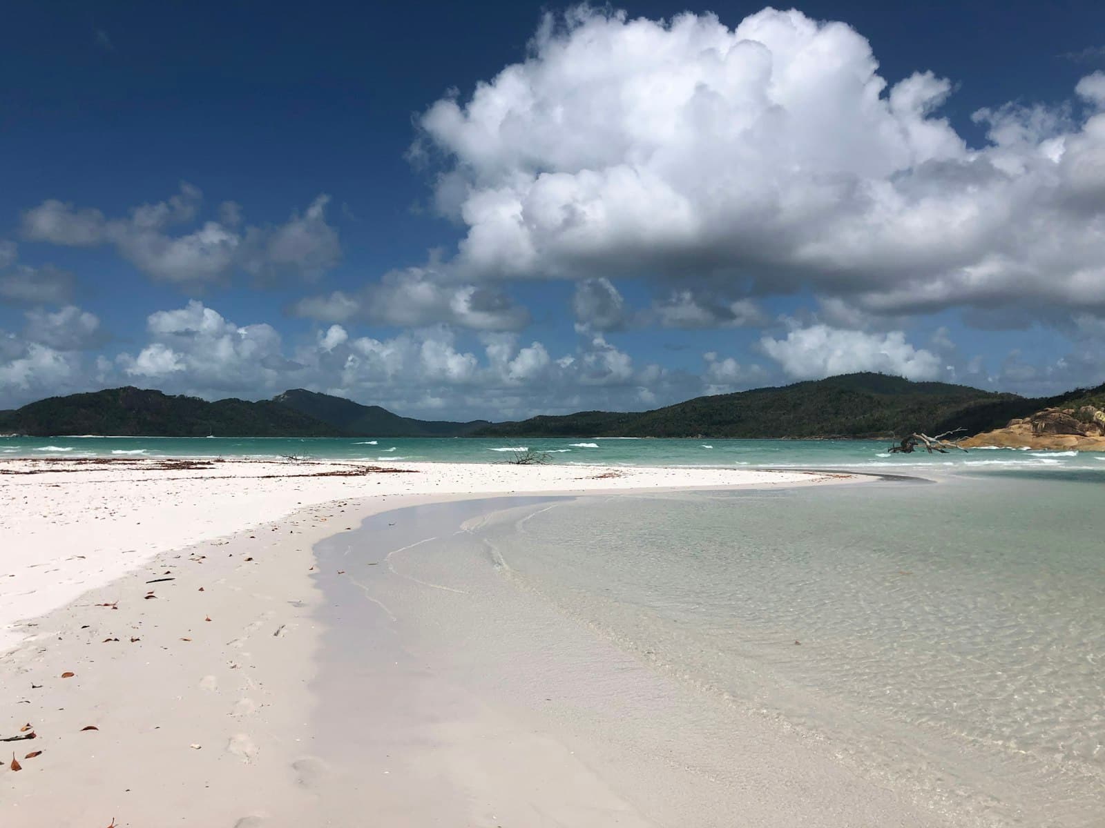 A pristine tropical beach with white sand, shallow turquoise waters, and a curved shoreline, backed by forested hills with granite outcrops visible on the right headland. - Photo by Pierre-Henry Soria on Unsplash