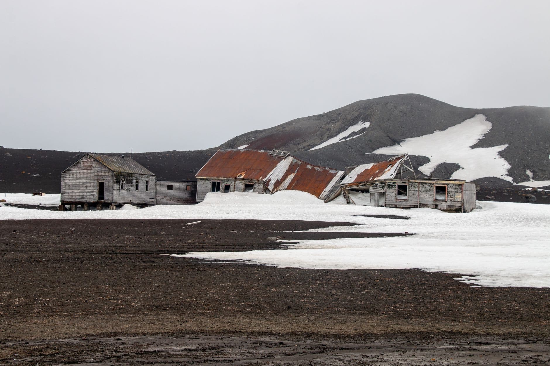 Derelict whaling station buildings on black volcanic sand with snow patches and a dark volcanic mountain in the background, showing the abandoned infrastructure at Whalers Bay, Deception Island. - Photo by ArcticDesire.com Polarreisen on Pexels