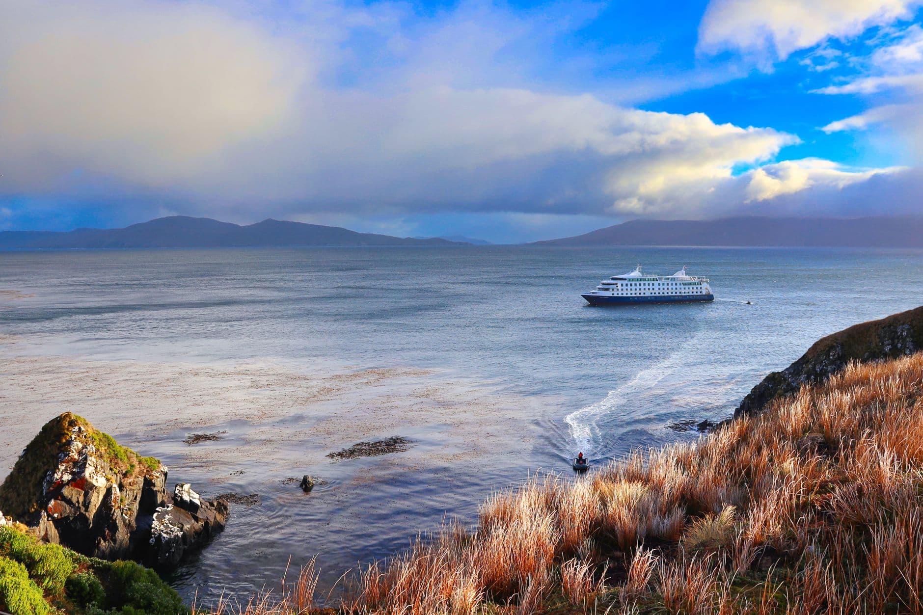 A modern expedition cruise ship anchored offshore in a Patagonian fjord, viewed from a rocky, grass-covered headland with mountains visible across the water under partly cloudy skies. - Photo by Dominik Ruhl on Pexels