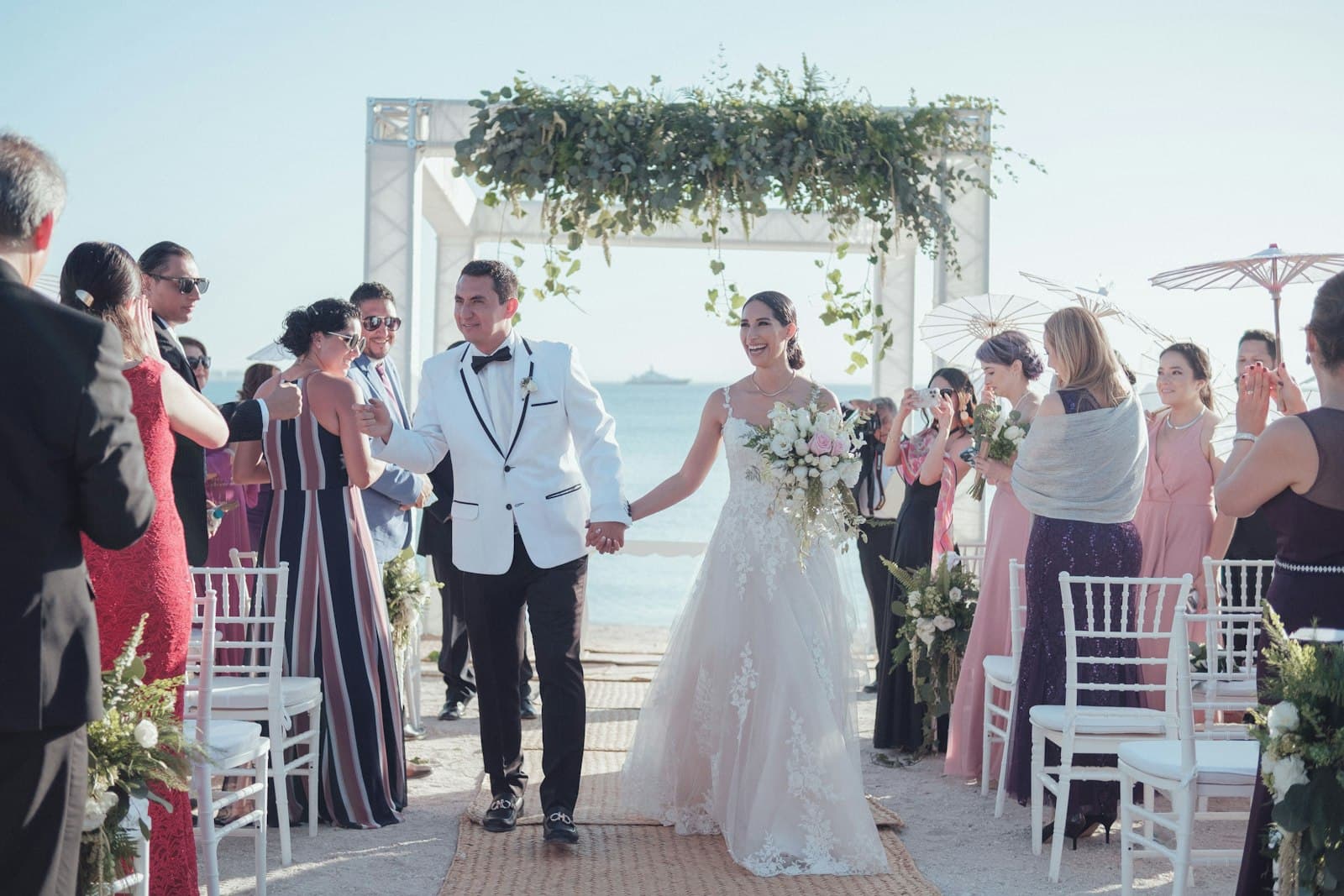 A beachfront wedding ceremony on a sandy beach overlooking the Caribbean Sea, with the bride and groom walking down the aisle under a decorated arch, surrounded by guests in formal attire. - Photo by Asdrubal luna on Unsplash