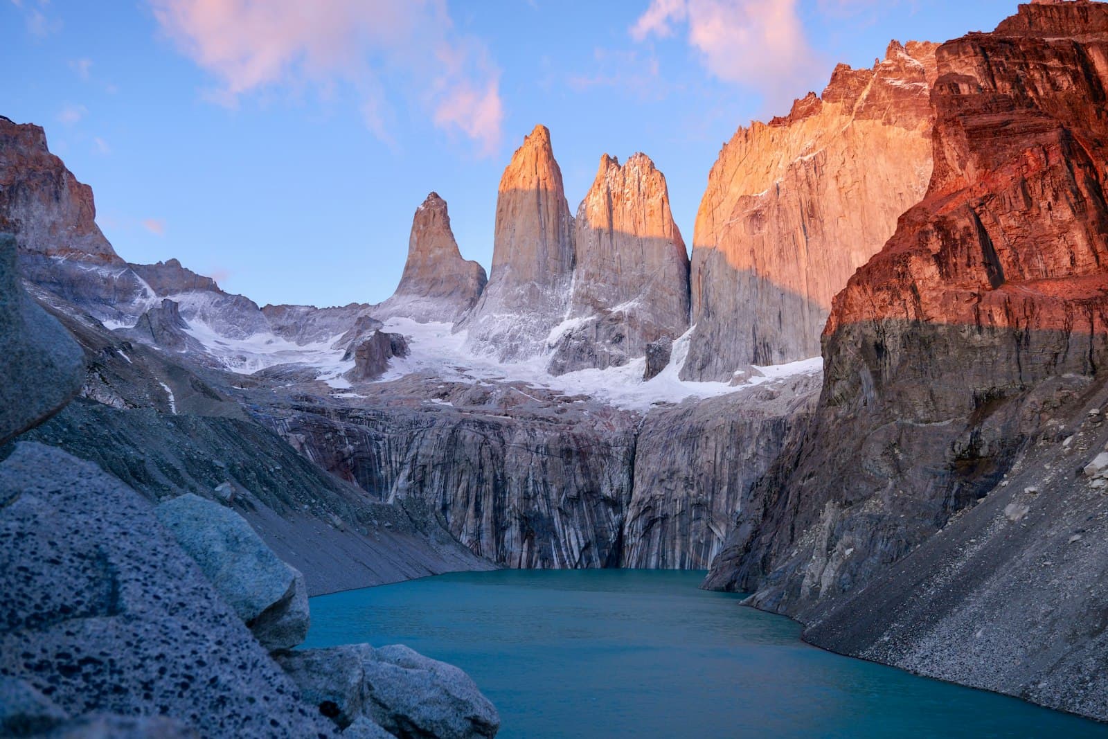 Dawn or dusk light illuminates three granite peaks rising above a turquoise glacial lake, with snow-covered slopes and columnar rock formations in a high-altitude Patagonian mountain valley. - Photo by Carter Obasohan on Unsplash
