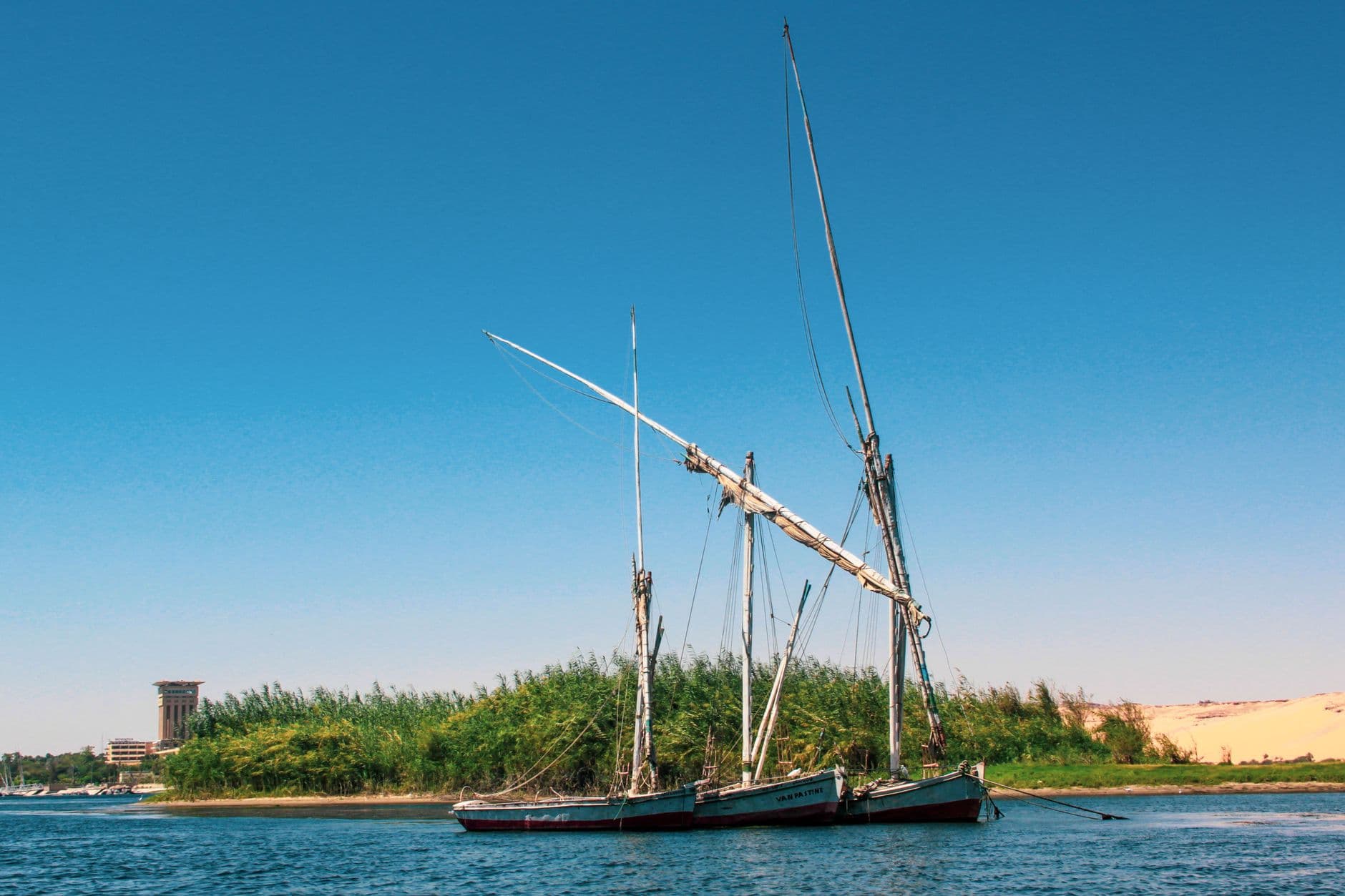 A traditional wooden dahabiya sailboat with two tall masts moored on the Nile River, with lush green riverbank vegetation and sandstone cliffs visible in the background under clear blue sky. - Photo by Yigithan Bal on Pexels