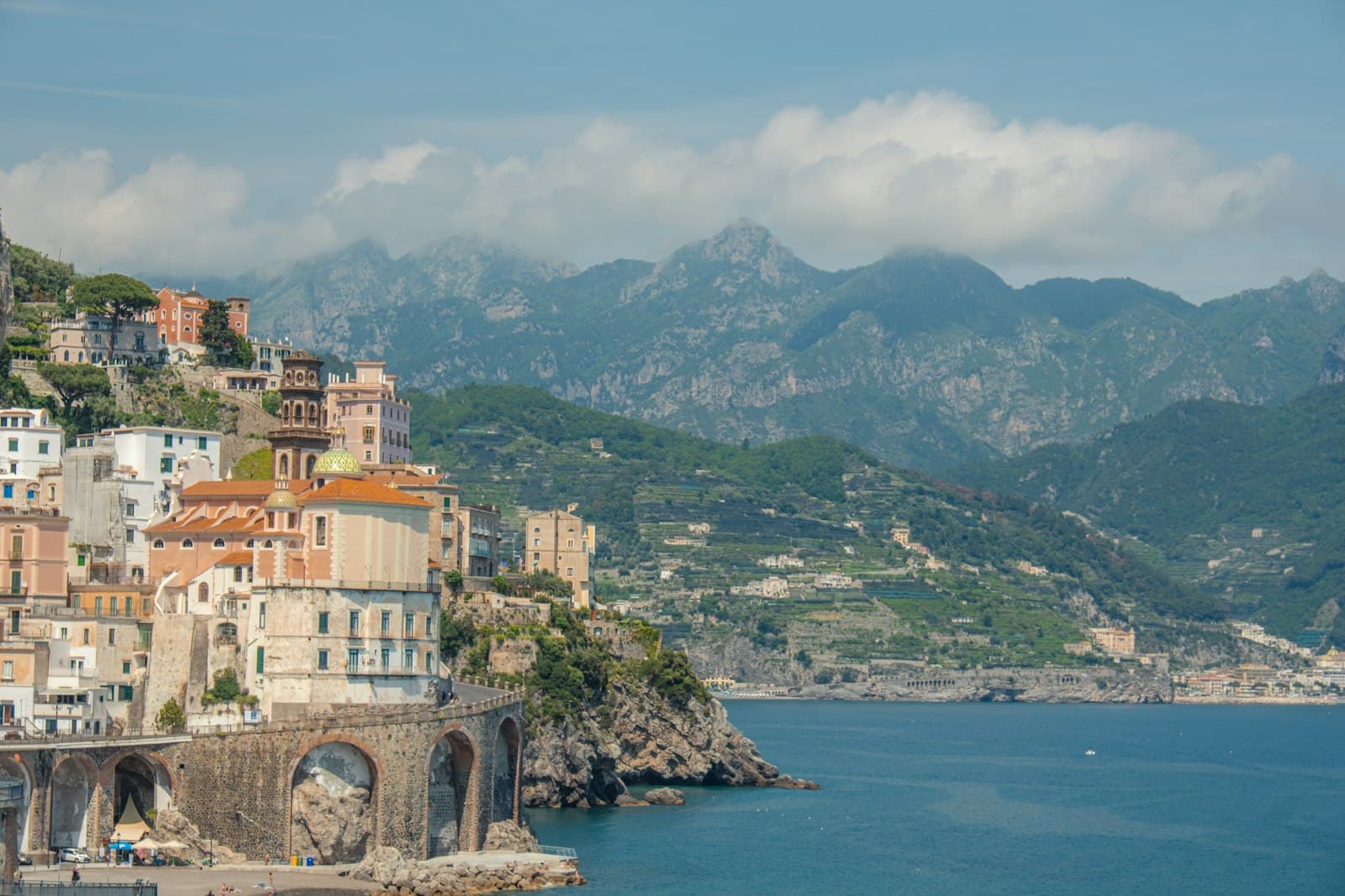 A picturesque clifftop village with pastel-hued buildings and orange-tiled roofs cascading down a rocky promontory above turquoise Mediterranean waters, with dramatic limestone mountains rising in the background. - Photo by Line Kjær on Unsplash