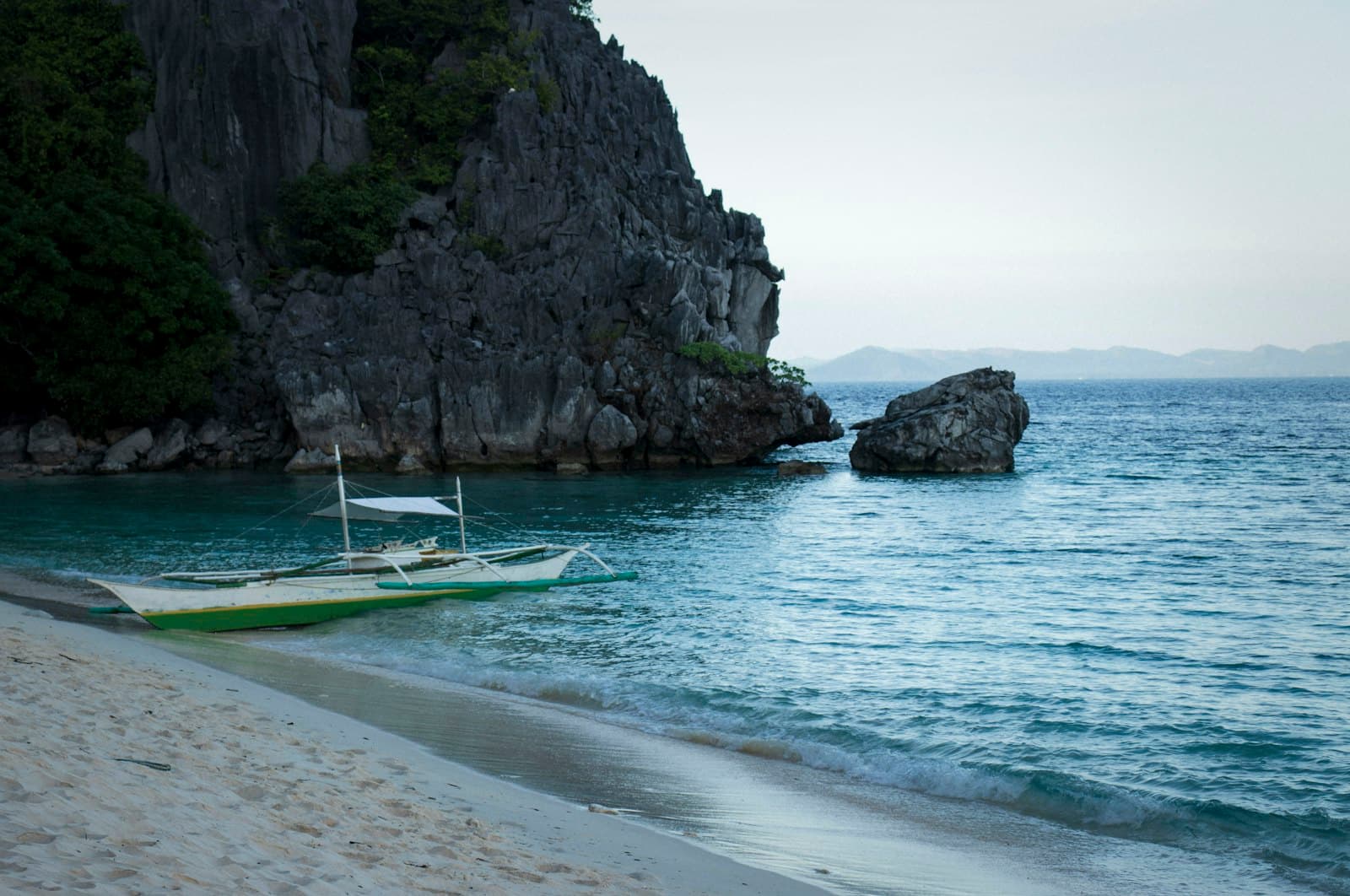 A traditional Philippine outrigger boat anchored in shallow turquoise water beneath dramatic limestone cliffs, with a pristine beach and distant islands visible across the bay. - Photo by Alaine Johnson on Unsplash