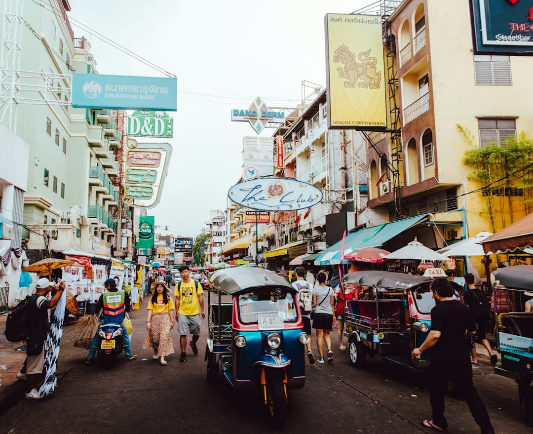 A busy Bangkok street market scene with crowds, street vendors, tuk-tuks, taxis, and commercial signage including visible Thai bank and business names. - Photo by Evan Krause on Unsplash
