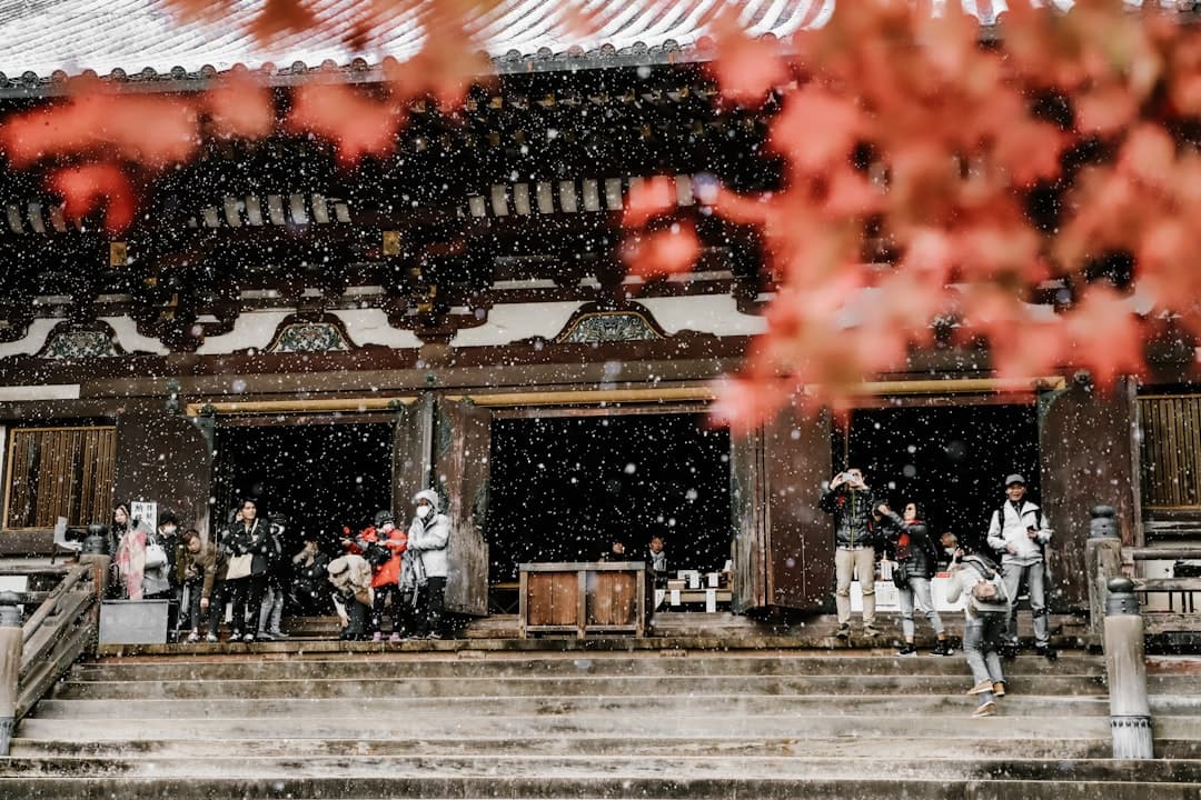 Snow falling on a traditional Japanese temple during what appears to be late winter or early spring, with visitors in winter clothing gathered on the temple grounds near blossoming cherry trees. - Photo by Steven Su on Unsplash