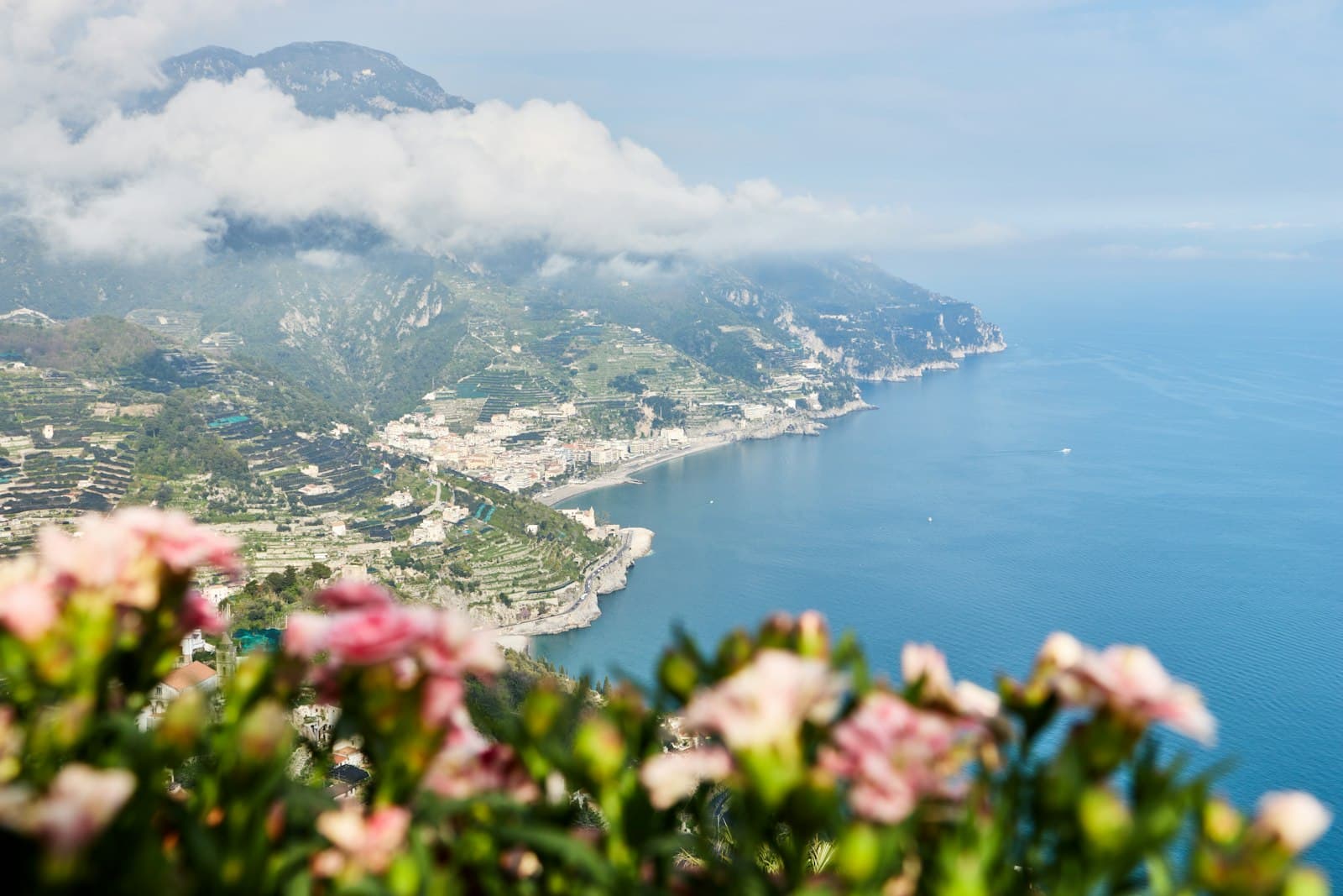 Aerial view of a coastal town nestled into steep cliffs on the Amalfi Coast, with pink flowers in foreground, terraced agricultural land, and deep blue Mediterranean Sea. - Photo by Matteo Eria on Unsplash
