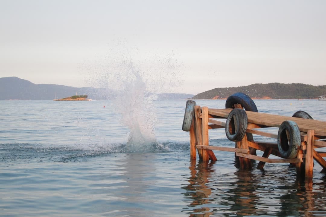 A wave crashes against the shore near weathered wooden structures with old tire bumpers, with forested islands visible across calm blue water under overcast sky. - Photo by Chris Linnett on Unsplash