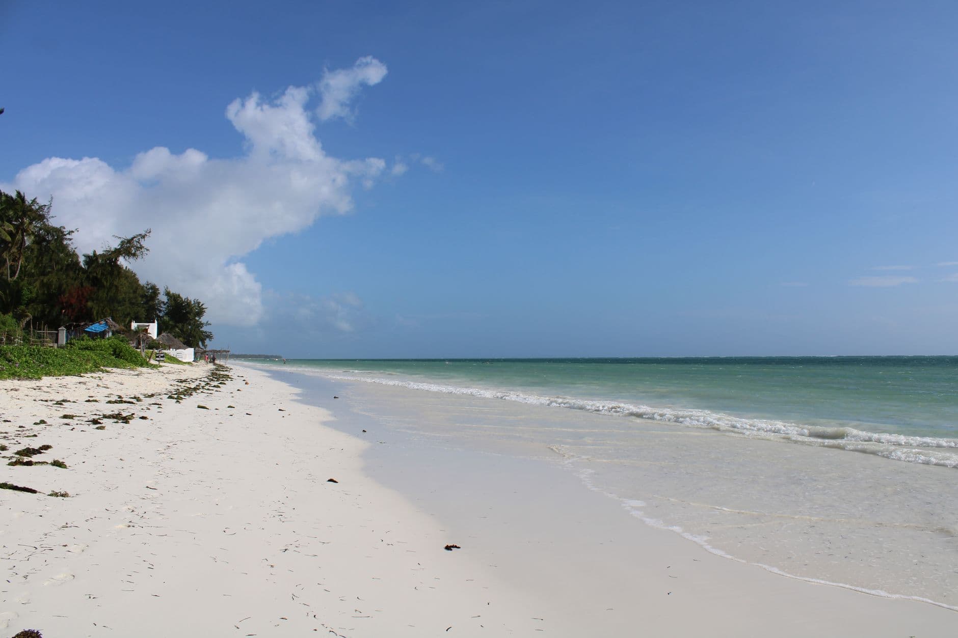 A pristine tropical beach with white sand, shallow turquoise waters, and gentle waves, with a small white building visible among vegetation on the left shore. - Photo by Ana Kenk on Pexels